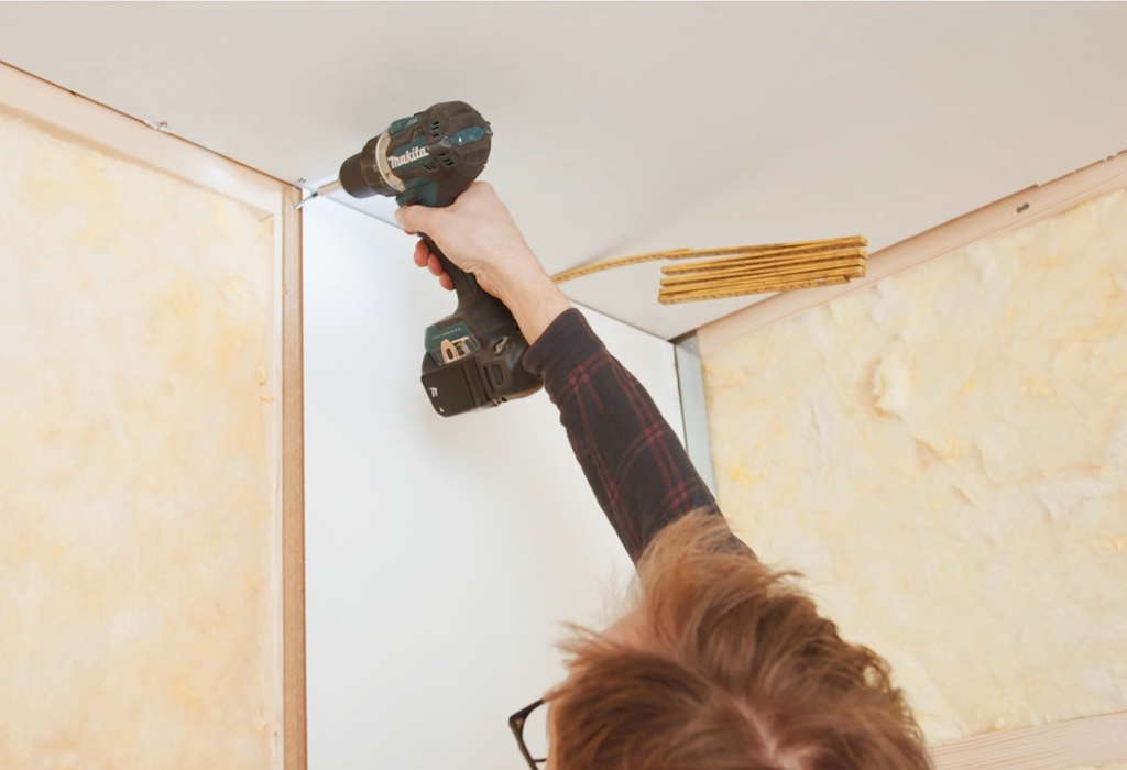 A person uses a cordless drill to attach a board to a wall in a room under construction, with insulation visible in the background.