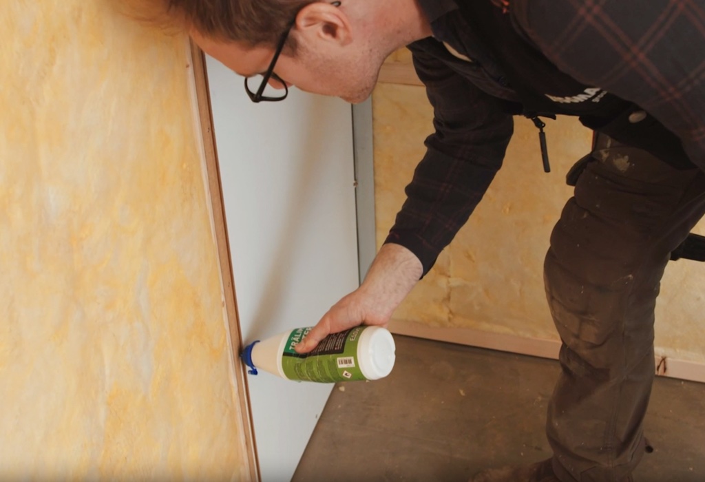 A person applies adhesive from a tube onto a panel indoors.