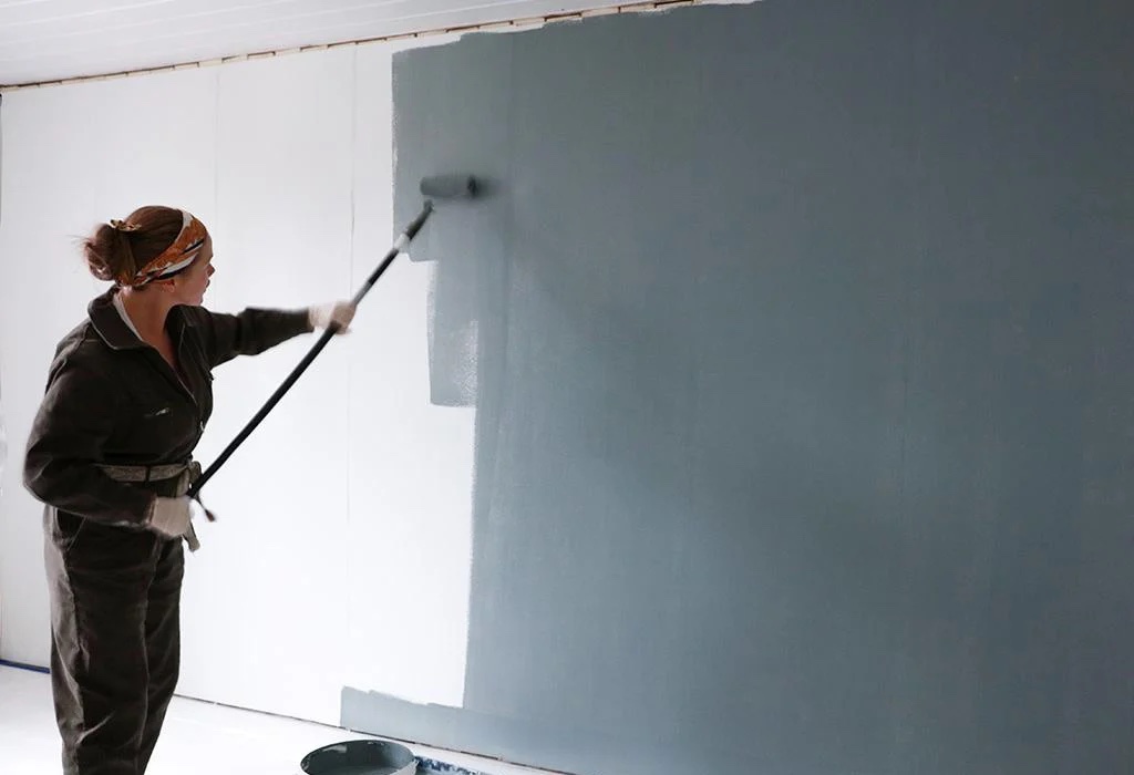 A person paints a white wall with a large roller brush, applying a coat of light blue paint.