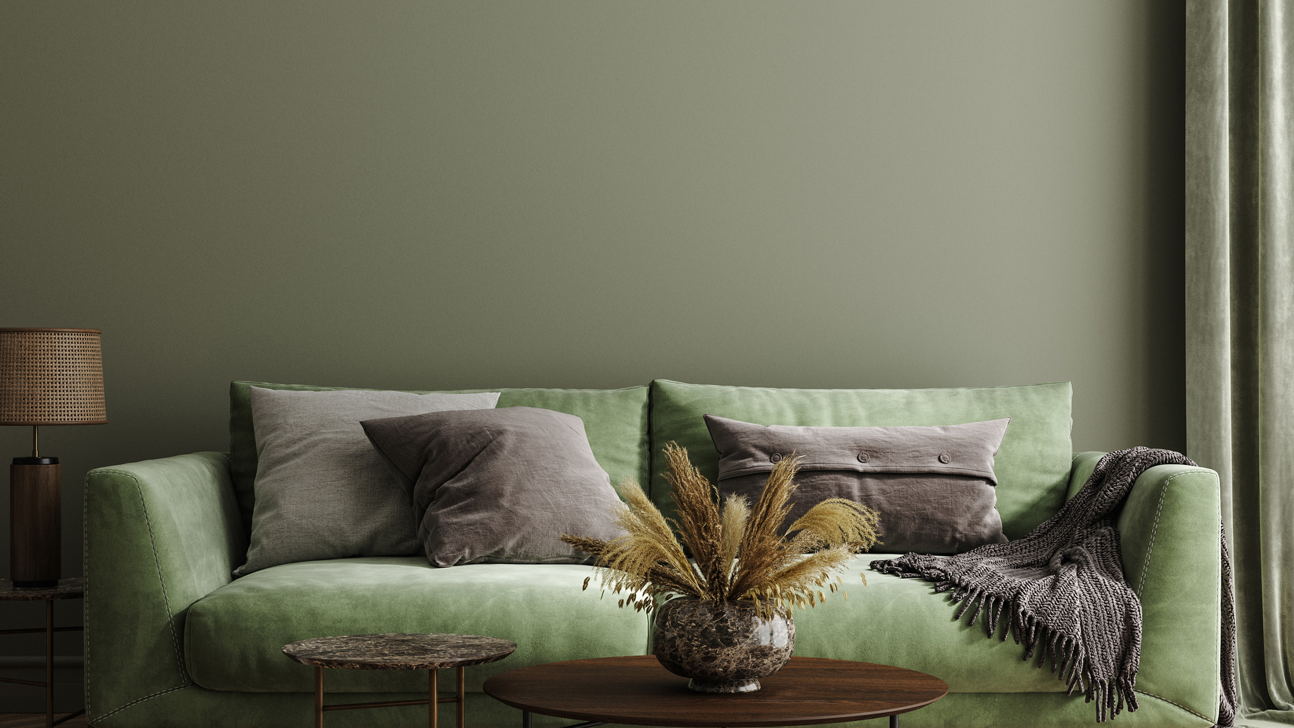 A green sofa with gray and beige pillows, a cozy throw, and a decorative vase with dried plants on a round wooden coffee table. A lamp is in the background.