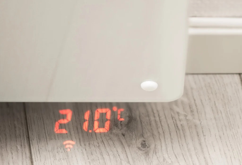 A digital display shows the temperature set at 21.0°C on a heater, with a wireless connectivity icon on a wooden floor.