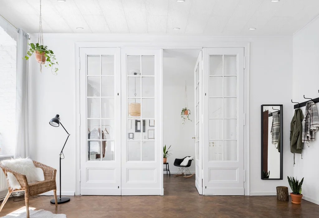 The bright room features large white French doors, a wicker chair, and a standing lamp. Coats hang near a full-length mirror on the right. Potted plants not only add greenery but also enhance indoor air quality, underscoring their importance in minimalist decor.