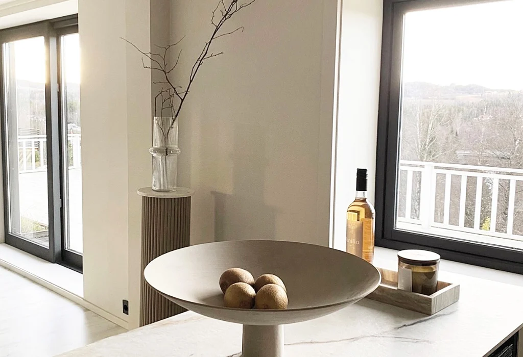 A modern kitchen with a window view showcases the importance of indoor air quality. A bowl with five apples sits on a counter, next to a bottle and small items on a tray, while a vase with branches enhances the ambiance of this well-appointed indoor environment.