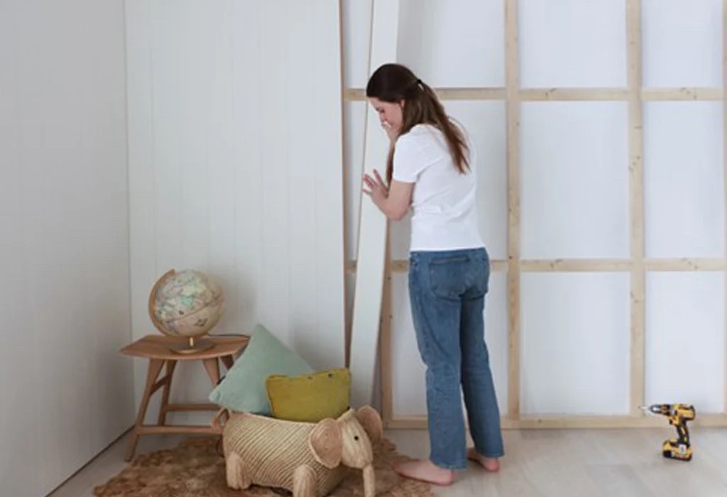 woman assembling a wooden wall panel in a room with a globe on a table, a woven elephant basket, and a drill on the floor.