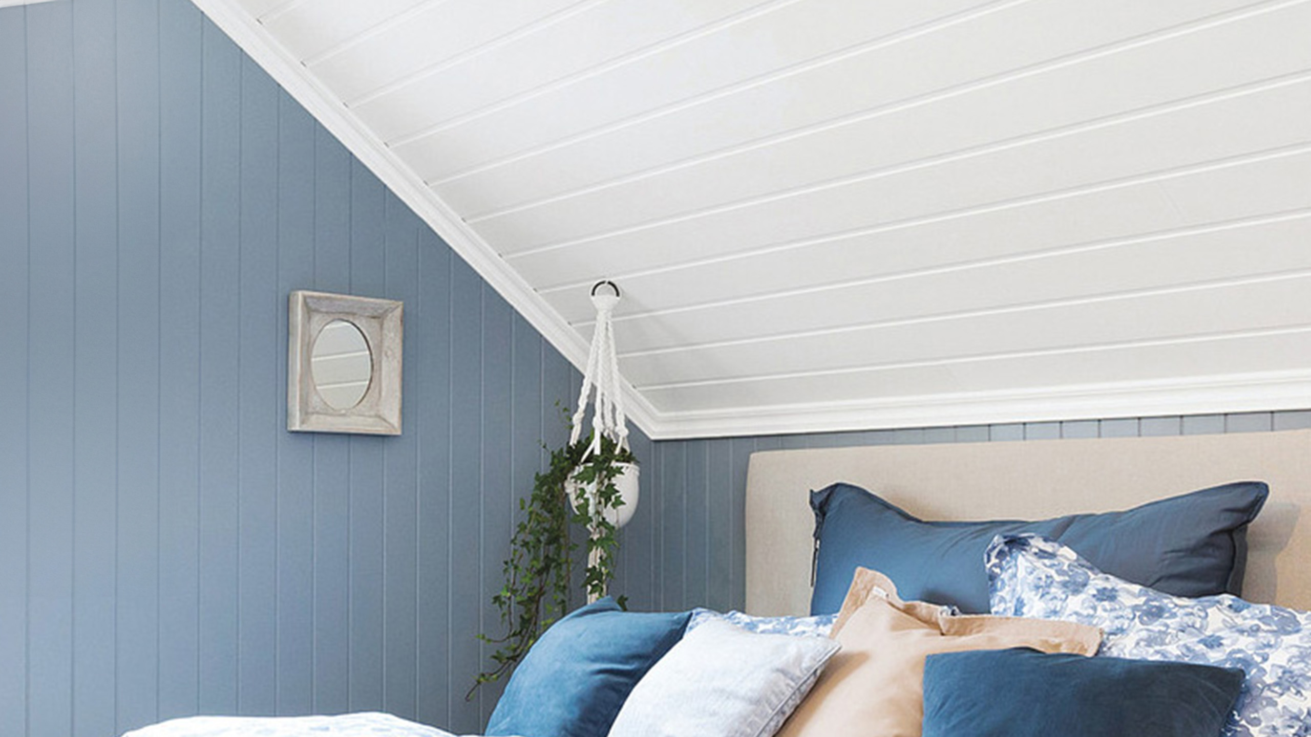 Bedroom with blue paneled walls, a white wood ceiling, and a bed with blue and white pillows. A small mirror and hanging decoration are on the wall.