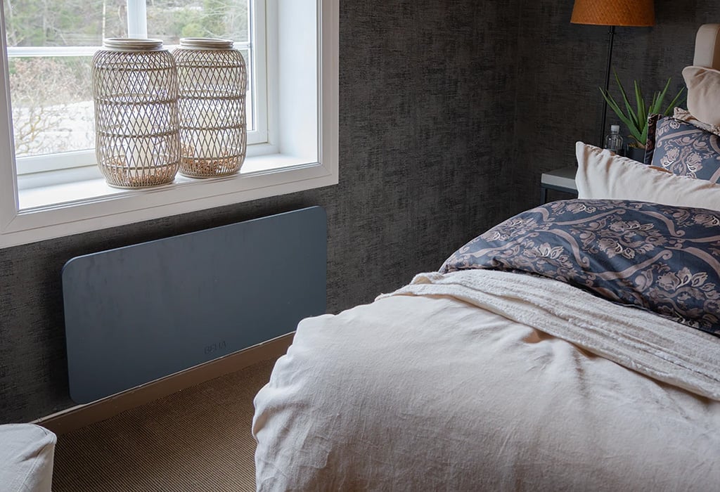 Bedroom with a bed, patterned blue and white bedding, a window, two wicker lanterns on the windowsill, a gray wall heater, and a lamp on a bedside table.